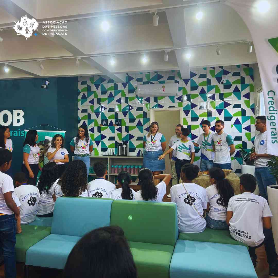 Na foto estão os alunos do curso interagindo com os colaboradores da agência, os alunos estão sentados num sofá na cor verde e azul e os colaboradores Sicoob em pé.